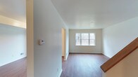 This two-floor space offers a straightforward and functional layout with a clean, modern aesthetic throughout. The main living area is finished with warm hardwood-style flooring that runs throughout, complementing the crisp white walls and creating a bright, welcoming atmosphere. A large multi-pane window allows generous natural light to fill the room, offering views of the surrounding wooded landscape beyond. The open flow of the main level is enhanced by a simple, uncluttered design that provides flexibility for a variety of furniture arrangements. A wall-mounted thermostat is conveniently positioned near the entryway, indicating climate control throughout the home. Archway openings connect the rooms smoothly, contributing to an easy, connected feel across the living spaces. The neutral color palette of soft whites and warm wood tones creates a calm and versatile interior that serves as a blank canvas for personalization. The second floor provides additional living space, further expanding the overall footprint of the home. Together, both levels offer a well-organized, move-in-ready environment suited for a range of lifestyles.