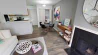 This well-appointed two-floor space showcases a thoughtfully designed interior that balances modern aesthetics with functional living. The main living and dining area features rich, dark wood-look flooring that runs continuously throughout, lending warmth and cohesion to the open-concept layout. Neutral gray walls provide a versatile backdrop, complemented by crisp white trim and accents that brighten the space.

The dining area is centrally positioned and accommodates a round glass-top table with sleek black chairs, creating an elegant yet casual setting for everyday meals. A built-in closet with louvered bifold doors adds practical storage nearby. Adjacent to the dining space, a wooden console table with open shelving serves as a functional accent piece, ideal for display and organization.

The kitchen flows seamlessly from the living area, featuring white cabinetry and a clean, bright aesthetic. The living area anchors the space with a defined seating zone, complete with a round coffee table set atop a textured area rug.

Throughout the space, carefully chosen decorative elements — including colorful artwork and curated accessories — add personality and visual interest. The overall ambiance is contemporary and welcoming, making the most of its open floor plan across two levels.