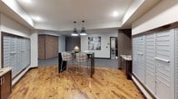 This well-appointed residential lobby combines modern design with practical functionality across a single floor. The space is anchored by rich, natural hardwood flooring that flows throughout the main gathering area, creating a warm and inviting atmosphere. A central high-top table with plaid upholstered bar-height chairs serves as a communal focal point, illuminated by a pair of striking industrial-style pendant lights that add a contemporary edge to the interior. Abstract artwork adorns the neutral-toned walls, complementing the clean lines and modern aesthetic of the space. A textured feature wall panel adds visual depth and contrast to the otherwise understated palette of grays and whites. Transitioning into the adjacent mail corridor, the flooring shifts to a durable gray carpet, maintaining a cohesive yet distinct functional zone. This area houses an extensive bank of individual mailboxes framed in dark wood trim, accompanied by a granite-topped cabinet for added convenience. The corridor itself is clean, well-lit, and efficiently laid out, with recessed lighting throughout ensuring the entire space remains bright. A glass-paneled door visible in the background hints at additional rooms or offices beyond. Overall, the lobby presents a polished, modern environment that balances community interaction with everyday residential needs.