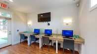 This Cyber Cafe is a thoughtfully designed, single-room technology hub built to provide a comfortable and functional environment for internet and computer access. The space features a long granite countertop workstation running along one wall, accommodating multiple desktop computers — each separated by clear glass privacy dividers — along with individual keyboards and mice, allowing several users to work simultaneously with a degree of privacy. Padded, ergonomic chairs are positioned at each station for comfortable extended use. A large flat-screen television is wall-mounted above the workstation area, flanked by two elegant wall sconces that provide warm, ambient lighting throughout the room. The flooring consists of rich wood-look planks that lend a clean, modern aesthetic to the space. Natural light filters in through transom-style windows near the ceiling and a full-lite glass entry door, which also offers a view of the surrounding outdoor greenery. White cabinetry beneath sections of the countertop provides additional storage. A printed informational sign is posted on the wall, offering guidance on cafe use. The layout is open and uncluttered, making the space easy to navigate and welcoming for all users.