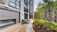 Located on Atlas Street in Baltimore's vibrant 21230 corridor, this outdoor amenity space is a carefully composed urban courtyard that blends natural landscaping with modern design. The space features a combination of warm wood decking and curved stone paver walkways that wind through lush planted garden beds filled with native shrubs, ornamental grasses, and slender young trees that provide greenery and shade.

Multiple distinct seating areas are distributed throughout the courtyard, offering a range of social settings. A round café table with chairs nestled against the brick building facade provides an intimate dining spot, while a central lounge arrangement of contemporary metal chairs surrounds a sleek circular fire bowl, creating an inviting gathering area. Pathway lighting is integrated at ground level throughout the garden beds, ensuring the space is functional and atmospheric after dark.

A dedicated outdoor kitchen and bar station, complete with a grill, sink, and bar-height seating, offers residents a fully equipped space for outdoor entertaining. Additional bench seating is positioned nearby for casual use. Black metal railing defines the perimeter, framing views of the surrounding Baltimore streetscape and neighboring brick architecture. The multi-story brick building facade, characterized by large industrial-style windows and Juliet balconies, forms an elegant backdrop to this well-appointed outdoor retreat.