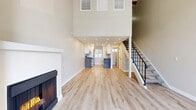 This two-story residence is thoughtfully designed across two floors of well-utilized living space. The main level immediately impresses with its dramatic double-height ceilings, creating an expansive, airy atmosphere in the living area. Light wood-tone flooring runs throughout the lower level, complementing the neutral palette of warm gray and white walls. A standout fireplace anchors the living room, while large shuttered windows and recessed lighting ensure the space remains bright and welcoming. The staircase, featuring a sleek black metal railing, adds a modern architectural element alongside the exposed wood beams visible overhead.

The kitchen is efficiently laid out with white shaker-style cabinetry, dark countertops, a gray subway tile backsplash, and stainless steel appliances, including a refrigerator and dishwasher. Natural light enters through a window overlooking the exterior.

Upstairs, the bedroom features exposed wood beam ceilings, plush carpet, and multiple plantation-shuttered windows that allow ample natural light. Sliding barn doors lead to the closet and bathroom areas, adding a stylish touch. Both bathrooms maintain a clean, modern aesthetic with white cabinetry, dark countertops, matte black hardware, and light hardwood-style flooring.