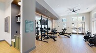 The Union Green Fitness Center at 4980 Stonewall Tell Rd in Union City, GA is a thoughtfully designed, single-floor exercise facility that offers a comprehensive workout environment. The space features light hardwood-style flooring throughout, crisp white walls, and high ceilings fitted with recessed lighting and ceiling fans that promote air circulation and a comfortable exercise atmosphere. Large windows along the perimeter flood the interior with abundant natural light and provide pleasant views of the surrounding greenery.

The facility is divided into distinct workout zones. The strength training area is anchored by a power rack with weight plates, a flat bench press, and a full dumbbell rack with adjustable benches, all set against a wall of mirrors that enhance the sense of openness. A dedicated functional training station houses kettlebells, battle ropes, medicine balls, resistance bands, and a BOSU balance trainer, catering to dynamic and cross-training workouts.

The cardio zone features multiple treadmills, an elliptical machine, a stationary bike, and a multi-function cable machine, all positioned near the windows for an energizing workout experience. Geometric linear pendant lighting accents the entry corridor, adding a modern architectural touch. The fitness center is also adjoined by a separate lounge space visible through glass-paneled doors, offering a seamless transition between active and social areas.