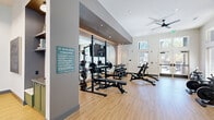 The Union Green Fitness Center at 4980 Stonewall Tell Rd in Union City, GA is a thoughtfully designed, single-floor exercise facility that offers a comprehensive workout environment. The space features light hardwood-style flooring throughout, crisp white walls, and high ceilings fitted with recessed lighting and ceiling fans that promote air circulation and a comfortable exercise atmosphere. Large windows along the perimeter flood the interior with abundant natural light and provide pleasant views of the surrounding greenery.

The facility is divided into distinct workout zones. The strength training area is anchored by a power rack with weight plates, a flat bench press, and a full dumbbell rack with adjustable benches, all set against a wall of mirrors that enhance the sense of openness. A dedicated functional training station houses kettlebells, battle ropes, medicine balls, resistance bands, and a BOSU balance trainer, catering to dynamic and cross-training workouts.

The cardio zone features multiple treadmills, an elliptical machine, a stationary bike, and a multi-function cable machine, all positioned near the windows for an energizing workout experience. Geometric linear pendant lighting accents the entry corridor, adding a modern architectural touch. The fitness center is also adjoined by a separate lounge space visible through glass-paneled doors, offering a seamless transition between active and social areas.