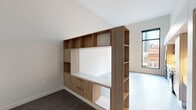 This contemporary single-floor residence showcases a thoughtfully designed open-concept layout that maximizes both functionality and aesthetic appeal. The space is characterized by soaring vaulted ceilings that amplify the sense of volume, while light hardwood-style flooring flows cohesively throughout the main living areas, transitioning to darker hardwood in the sleeping zone.

The kitchen is fully equipped with a stainless steel suite of appliances — including a full-sized refrigerator, dishwasher, gas range, and microwave — complemented by light wood cabinetry with under-cabinet lighting and clean white countertops. Adjacent to the kitchen, a built-in desk area provides a dedicated workspace seamlessly integrated into the cabinetry design.

A standout feature of the living area is a freestanding custom wood shelving unit that serves as both a functional storage wall and a design centerpiece, complete with open shelves, closed cabinetry, and a built-in media niche. The unit doubles as a room divider, adding architectural interest without sacrificing openness.

Large, black-framed floor-to-ceiling windows frame sweeping urban skyline views, flooding the interior with natural light and connecting the space to its vibrant city surroundings. A smart thermostat panel is also present, reflecting the residence's modern, tech-integrated conveniences. The overall ambiance is clean, minimalist, and sophisticated.