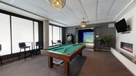 Located at 3250 W 66th St in Edina, Minnesota, this single-floor amenity lounge offers a well-appointed recreational space designed for socializing and entertainment. The layout flows seamlessly from a welcoming lounge entry — furnished with curved, upholstered accent chairs flanked by warm wall sconces and a subtly textured feature wall — into a larger open activity area. At the heart of the space sits a full-size billiards table set atop rich herringbone-patterned hardwood flooring, creating a natural focal point. Adjacent to the billiards area, bar-height tables and stools are arranged in front of a large picture window, offering an ideal setting for casual conversation or work. Pendant lighting in varied styles adds character throughout, while the dark accent walls and neutral gray tones provide a contemporary, sophisticated ambiance. A dedicated Full Swing golf simulator room occupies one end of the space, enclosed in dark paneling with turf flooring and a large projection screen, providing an immersive experience for golf enthusiasts. Decorative touches, including framed artwork and an arched display cabinet, contribute warmth and personality to the overall design. The space connects to adjoining areas through glass-panel doors, maintaining an open and airy feel throughout.