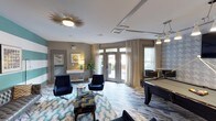 Located at 712 Tucker St in Raleigh, NC, this single-floor community clubhouse delivers a polished, contemporary atmosphere through thoughtful design and upscale finishes. The main lounge area serves as the centerpiece of the space, anchored by a striking sunburst ceiling fixture and accented by geometric tray ceiling details. Plush navy blue velvet swivel chairs and a bold, wave-patterned area rug create an inviting seating arrangement over marble-look tile flooring that runs throughout. A full-size billiards table occupies a dedicated game area, complete with overhead pendant lighting and a large wall-mounted television, flanked by houndstooth-patterned accent wallpaper that adds visual depth. Floor-to-ceiling glass French doors flood the space with natural light and open directly onto an outdoor patio, offering views of an exterior grilling and lounge area. A compact, well-appointed kitchenette features dark espresso cabinetry, granite countertops, a subway tile backsplash, and stainless steel appliances. A separate work and business area provides a dedicated desk with computer access, set against a striking teal and white striped feature wall. The overall design balances functionality with mid-century modern aesthetics, making it a versatile and welcoming communal destination.