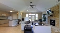 This single-floor space is designed with an open, airy feel accentuated by vaulted ceilings that add a sense of generous vertical space throughout the main living area. A black ceiling fan is centrally mounted, providing both function and a subtle design accent against the smooth, angled ceiling. Recessed lighting complements the abundant natural light streaming in through a set of French doors flanked by large windows along the back wall, offering views to the outdoors.

The living area is furnished with neutral-toned sofas and accent chairs arranged around a dark-finished coffee table, creating a cohesive and inviting seating arrangement. Decorative throw pillows in muted patterns add visual interest without overwhelming the calm palette. Light beige tile flooring runs throughout, enhancing the bright, clean aesthetic of the space.

To one side, a raised bar counter with tall bar-style seating connects to an adjacent area, suggesting a kitchen or dining space nearby. Wall sconces and a large round clock add decorative touches to the neutral-painted walls. A tall potted plant in the corner introduces a natural element, contributing to the overall warm and welcoming ambiance of the space.