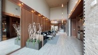 This single-story lobby showcases a striking contemporary design defined by soaring double-height ceilings that lend an expansive, airy quality to the space. Warm wood-paneled walls line the interior, providing a rich, natural contrast against the light-toned large-format tile flooring laid in a subtle herringbone pattern. Pendant bulb lighting descends from above on varied-length cords, casting a soft, ambient glow throughout the space.

A built-in lounge area features deep charcoal seating accented with patterned throw pillows, accompanied by sleek gold-legged side tables arranged atop a gray area rug. Decorative oversized planters display sculptural bare-branch arrangements alongside low succulent plantings, adding organic texture and visual interest to the environment.

On the opposite side, a bold black-and-white geometric column or wall feature provides a striking graphic accent, balancing the warmth of the wood tones. The far end of the lobby opens toward glazed doors, allowing natural light to filter through and providing a sense of depth. Overall, the space strikes a refined balance between natural warmth and modern minimalism.