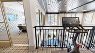 Located at The Enclave on Piney Lane in Asheville, NC, this single-floor amenity space offers a diverse range of facilities designed for comfort, productivity, and wellness. The fitness area is a standout feature, equipped with cycling bikes, an upright exercise machine, and a treadmill, all set against a backdrop of stylish built-in shelving displaying decorative accents and curated artwork. A striking natural stone accent wall and exposed beam ceiling add texture and warmth, while large windows flood the room with natural light and offer views of the surrounding landscape. Plaid carpeting runs throughout, tying the space together with a cohesive aesthetic. Adjacent to the fitness area, a dedicated conference room provides a polished setting for meetings, featuring a solid wood communal table, upholstered rolling chairs, geometric patterned wallcovering, pendant lighting, and a wall-mounted display screen. Floor-to-ceiling windows along one wall invite abundant daylight and outdoor views. A pet grooming room rounds out the amenities, outfitted with a utility sink, green cabinetry, decorative tile, wall-mounted shelving with pet-themed décor, and a cozy seating area — all finished with clean white walls and a welcoming, functional layout.