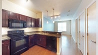 This single-floor space is designed with an open-concept layout that seamlessly connects the kitchen and living area. The kitchen is a standout feature, showcasing rich dark wood cabinetry paired with dark granite countertops that extend into a peninsula, offering both additional prep space and a natural divider between the kitchen and the adjoining living area. Black appliances, including a range, built-in microwave, and dishwasher, complement the cabinetry for a cohesive, contemporary look. A double-basin sink is positioned along the peninsula, adding functional convenience. Overhead, a track lighting fixture with pendant lights illuminates the kitchen, creating a warm and inviting atmosphere. The flooring throughout is a warm hardwood-style material that adds continuity and a sense of flow across the space. The living area benefits from a window that allows natural light to filter in, brightening the room and enhancing the open feel. Soft, neutral wall tones in a muted blue-green complement the warm wood tones throughout. A hallway visible beyond the living area suggests additional rooms, contributing to the overall functionality of this well-appointed space.
