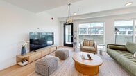 Located at 6019 Dunbar Street in Vancouver's desirable Dunbar neighbourhood, this single-floor rooftop amenity space offers a well-appointed interior complemented by an expansive outdoor terrace. The interior is divided into a combined dining and living area connected by a hallway, finished with light hardwood flooring and crisp white walls that create a clean, contemporary aesthetic. The dining area features large sliding glass doors and floor-to-ceiling windows that flood the space with abundant natural light and frame views of the surrounding neighbourhood and sky. A stylish pendant light fixture anchors the dining space, which comfortably accommodates a full dining table and chairs. The adjacent living area is equally bright, with sliding glass doors providing direct rooftop access, a wall-mounted display, seating, and a pendant chandelier that adds a warm focal point. The outdoor terrace is a standout feature, finished with large-format porcelain tile pavers and equipped with built-in planters, pergola structures, and bench seating. A dedicated play area with colourful rubber surfacing and sculptural elements adds a lively, community-oriented character. The exterior facade presents a modern composition of panel cladding with warm timber accents, and the terrace enjoys open sky views with surrounding tree canopies visible on the horizon.