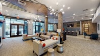 The clubhouse at Sterling Apartment Homes in Smyrna, Tennessee, is a thoughtfully designed community facility that combines contemporary aesthetics with functional gathering spaces. The entrance greets visitors with large glass doors, marble-look tile flooring, and an immediate sense of openness and light.

The main lounge area is expansive and inviting, featuring multiple seating arrangements with neutral-toned sofas accented by bold orange and blue throw pillows. A striking wood-paneled ceiling detail with angular geometric lines draws the eye upward, complemented by sculptural sputnik-style chandeliers that add a modern flair. A linear fireplace and wall-mounted television anchor one side of the lounge, while floor-to-ceiling windows flood the space with natural light and provide views of the surrounding exterior.

A stylish bar area features a long stone-topped counter with upholstered bar seating, a textured blue-gray tile backsplash, and pendant lighting overhead. Adjacent dining nooks with round wood tables and leather chairs offer additional casual seating near large windows overlooking an outdoor courtyard.

A secondary lounge area near the entrance showcases rich cognac-colored accent chairs, bold artwork, and a coffee station, creating an energetic and welcoming atmosphere. A staircase with wood paneling and industrial metal railings serves as a dramatic architectural focal point throughout the space.