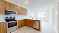 This thoughtfully designed single-floor space showcases a clean, contemporary aesthetic throughout. The open-concept kitchen is a standout feature, boasting rich, warm-toned wood grain cabinetry — both upper and lower — complemented by white quartz countertops and a patterned white tile backsplash. Stainless steel appliances, including a range, built-in microwave, and dishwasher, add a polished, functional edge. A peninsula with matching wood paneling extends into the living area, creating a natural division of space while maintaining an open, airy flow. Light hardwood flooring runs consistently throughout, enhancing the brightness of the white walls and recessed lighting overhead.

The bedroom offers a generous picture window that frames views of the surrounding neighborhood, flooding the room with natural light, while a ceiling fan adds comfort year-round. The space includes two bathrooms: one features a full soaking tub surrounded by classic white subway tile and a floating vanity with a wood-accented base, while the second presents marble-look tile flooring, a floating white vanity, and a sleek glass-enclosed shower. Together, the interiors reflect a cohesive modern design with attention to detail and quality finishes throughout.