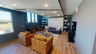 Located in Northlake, TX, this single-floor community amenity space is thoughtfully designed to foster social interaction and relaxation. The interior is anchored by bold teal and deep navy accent walls that create a vibrant yet cohesive atmosphere, complemented by warm wood-look flooring that runs throughout the entire space.

The main lounge area features a plush navy plaid sofa accented with yellow and patterned throw pillows, flanked by a pair of rich brown barrel chairs. A striking focal wall of white textured tile panels is framed by teal built-in open shelving on both sides, tastefully styled with books, decorative objects, and accent pieces. Bold gold-and-white patterned armchairs surround a glass-top coffee table, providing additional seating for gatherings.

The game room section includes a full-size pool table positioned beneath a coffered ceiling with recessed lighting, alongside a wall-mounted television, making it an ideal space for entertainment. Tall vertical windows throughout the space flood the interior with natural light and offer views of the surrounding Northlake landscape.

A smaller nook features a round wood dining table with metal chairs beneath a brass pendant light, built-in shelving, and a decorative arrangement of circular gold-framed mirrors. A glass-panel door connects to an adjacent fitness area, enhancing the overall functionality of this well-appointed community space.