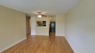 This single-story space features an open-concept living area finished with warm hardwood-style flooring that runs throughout, creating a cohesive and inviting atmosphere. The living area is spacious and unobstructed, benefiting from a ceiling fan with a built-in light fixture that provides both air circulation and overhead illumination. A textured popcorn ceiling adds a classic residential character to the space.

The kitchen is accessible via a pass-through cutout in the wall, offering a convenient connection between the cooking and living areas. It is equipped with black appliances, including a refrigerator and microwave, complemented by warm wood-toned cabinetry and what appears to be a dark countertop surface. A sink is positioned beneath the pass-through window, adding practicality to the layout.

A hallway visible along the left side of the living area suggests additional rooms beyond, providing further functional space. The neutral beige wall tones throughout contribute to a clean, understated aesthetic that is versatile and easy to personalize. Overall, the layout is practical and well-connected, suited for everyday living.