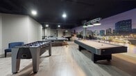 Located at 100 Trumbull in Hartford, Connecticut, this single-floor game room is a well-appointed recreational space designed for entertainment and leisure. The room is defined by a striking matte black ceiling that creates a bold contrast against the light wood-plank flooring, lending the space a sleek, modern aesthetic. A large, panoramic wall mural depicting the Hartford city skyline at dusk serves as the room's dramatic focal point, adding a sense of local character and visual depth.

The space is equipped with multiple gaming stations, including a full-size pool table illuminated by classic green pendant lights, a foosball table, and an air hockey table, offering a diverse range of recreational activities. A large floor-to-ceiling mirror along one wall visually expands the room and enhances its open feel. Recessed ceiling lighting provides consistent, even illumination throughout the space, complementing the pendant fixtures above the pool table.

A designated lounge area furnished with chairs and bar-height stools offers comfortable seating for spectators or those taking a break from the games. The overall layout is open and spacious, allowing for easy movement between activity areas. The combination of entertainment options, cohesive dark-and-light design palette, and the distinctive skyline mural makes this game room a dynamic and inviting communal space.