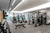 This contemporary fitness center is thoughtfully designed to accommodate a full range of workout styles across its single-level layout. The main exercise floor is expansive, finished with durable rubber flooring, and outfitted with a comprehensive selection of cardio equipment including ellipticals, StairMaster machines, and additional cardiovascular units positioned along floor-to-ceiling windows that frame outdoor views and draw in natural light. Recessed cove lighting along the ceiling perimeter creates a warm, modern ambiance throughout.

A dedicated free weights section features an extensive dumbbell rack spanning a wide range of weights, multiple adjustable benches, kettlebells, and cable machines, all set against mirrored walls that enhance the sense of space. A large flat-screen television is mounted nearby for added convenience.

A separate studio room offers a distinctly different atmosphere, featuring light hardwood flooring, full-length mirrors, mounted televisions, medicine ball storage, and yoga mats — ideal for stretching, yoga, or group fitness sessions. Glass partition walls and views to an outdoor courtyard give the studio an open, airy feel. The overall design is sleek and modern, with a dark and neutral color palette punctuated by bold teal accent walls.
