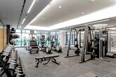 This contemporary fitness center is thoughtfully designed to accommodate a full range of workout styles across its single-level layout. The main exercise floor is expansive, finished with durable rubber flooring, and outfitted with a comprehensive selection of cardio equipment including ellipticals, StairMaster machines, and additional cardiovascular units positioned along floor-to-ceiling windows that frame outdoor views and draw in natural light. Recessed cove lighting along the ceiling perimeter creates a warm, modern ambiance throughout.

A dedicated free weights section features an extensive dumbbell rack spanning a wide range of weights, multiple adjustable benches, kettlebells, and cable machines, all set against mirrored walls that enhance the sense of space. A large flat-screen television is mounted nearby for added convenience.

A separate studio room offers a distinctly different atmosphere, featuring light hardwood flooring, full-length mirrors, mounted televisions, medicine ball storage, and yoga mats — ideal for stretching, yoga, or group fitness sessions. Glass partition walls and views to an outdoor courtyard give the studio an open, airy feel. The overall design is sleek and modern, with a dark and neutral color palette punctuated by bold teal accent walls.