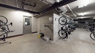 This indoor bicycle storage facility is a purpose-built space designed to accommodate a large number of bikes in an organized and secure environment. The room features a combination of freestanding floor-mounted racks and wall-mounted hooks, allowing bicycles to be stored both vertically and horizontally, maximizing the available floor space. The layout accommodates a wide variety of bicycle types, from road bikes to mountain bikes and folding models.

The space has a utilitarian, industrial aesthetic, with exposed concrete walls and a polished concrete floor that is easy to maintain. Overhead, exposed pipes, ductwork, and fluorescent strip lighting run along the ceiling, contributing to the no-frills, functional character of the room. The lighting provides adequate illumination throughout the space.

Access to the storage area is provided through a secured door with a window, ensuring controlled entry. Additional amenities include a wall-mounted bicycle repair station, offering basic tools for on-site maintenance, along with what appears to be an air compressor for tire inflation. This well-equipped storage room reflects a strong commitment to cyclist convenience and sustainability.
