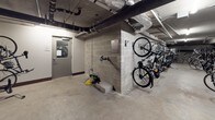 This indoor bicycle storage facility is a purpose-built space designed to accommodate a large number of bikes in an organized and secure environment. The room features a combination of freestanding floor-mounted racks and wall-mounted hooks, allowing bicycles to be stored both vertically and horizontally, maximizing the available floor space. The layout accommodates a wide variety of bicycle types, from road bikes to mountain bikes and folding models.

The space has a utilitarian, industrial aesthetic, with exposed concrete walls and a polished concrete floor that is easy to maintain. Overhead, exposed pipes, ductwork, and fluorescent strip lighting run along the ceiling, contributing to the no-frills, functional character of the room. The lighting provides adequate illumination throughout the space.

Access to the storage area is provided through a secured door with a window, ensuring controlled entry. Additional amenities include a wall-mounted bicycle repair station, offering basic tools for on-site maintenance, along with what appears to be an air compressor for tire inflation. This well-equipped storage room reflects a strong commitment to cyclist convenience and sustainability.
