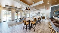 Located at 2100 E Aventura Way in Maryland Heights, Missouri, this single-story community clubhouse offers a well-appointed and thoughtfully designed amenity space. The welcoming entrance greets visitors with soaring ceilings, a striking geometric chandelier, textured stone accent walls, and wood-plank flooring that flows throughout the facility.

The main gathering area is an open, light-filled lounge and dining space featuring coffered ceilings with warm wood inlays, industrial-style pendant lighting, and expansive windows that frame views of the outdoor pool area. High-top tables with bar seating blend seamlessly with a comfortable lounge arrangement, creating a versatile social environment.

The kitchen and bar area is outfitted with natural wood cabinetry, white quartz countertops, a coffee station, and a wall-mounted television — accented by warm pendant lights and plantation shutters for a refined, rustic aesthetic. A separate, moodier dining nook with rich wallcoverings and table seating provides an intimate gathering spot.

The fitness facilities are split across two dedicated exercise rooms. One features a full lineup of cardio machines, mirrored walls, large windows overlooking the grounds, and ceiling fans. The second is equipped for strength and cycling training, with free weights, resistance equipment, and spin bikes.

A private office workstation with a computer and printer rounds out the offerings, providing a functional workspace within this comprehensive community hub.