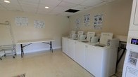 This single-level shared laundry facility is designed to provide residents with a functional and straightforward space for their laundry needs. The room features a row of white top-load washing machines and dryers aligned along one wall, offering ample capacity for multiple users. Instructional posters — including guides for easy washing and simple drying — are mounted above the machines, making the space accessible and user-friendly for all residents.

A folding table is positioned along the opposite wall, providing a dedicated surface for sorting and folding laundry. The light-toned walls and tile flooring create a clean, neutral environment that is easy to maintain. Recessed lighting in the drop ceiling ensures adequate illumination throughout the room.

The layout is open and efficiently organized, maximizing usable floor space while keeping the area uncluttered. Overall, this laundry facility prioritizes practicality and ease of use, serving as a convenient shared amenity within the community.