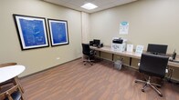 This single-room business center is designed to support a range of professional tasks in a clean, functional environment. The space features warm wood-look flooring, a drop ceiling with recessed panel lighting, and neutral sage-toned walls that create a calm, focused atmosphere.

Along one wall, a continuous built-in countertop workstation accommodates multiple desktop computers with ergonomic rolling chairs, providing ample workspace for individual use. A multifunction device offering print, scan, copy, and fax services is conveniently stationed along the same counter, making it easy to handle a variety of document needs in one location. Framed blueprint-style artwork adds a touch of understated character to the walls.

A small round table with wooden chairs occupies a corner of the room, offering a casual spot for brief meetings or collaborative work. Posted business center rules ensure clear guidelines for users of the space.

Access is provided through a gray door with a narrow window panel, maintaining privacy while allowing easy entry. The overall layout is efficient and well-organized, catering to guests or tenants who require reliable office amenities without sacrificing comfort. This business center delivers a practical and professional setting for productivity.
