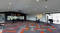 Located at 800 Baldwin Dr in Grandview Heights, Ohio, this fitness center offers a well-appointed, single-floor workout facility designed to motivate and accommodate a range of exercise needs. The space is immediately striking thanks to its bold, geometric carpet in a vibrant pattern of orange, gray, and charcoal — a dynamic visual element that energizes the entire floor. White walls contrast with a dark accent wall, giving the space a modern, high-energy aesthetic that is both functional and visually appealing. Recessed lighting throughout the ceiling ensures the space is well-lit, while ceiling fans provide comfortable airflow during workouts. A large wall mirror on the accent wall expands the sense of space and allows exercisers to monitor their form. The facility is equipped with strength training machines, a dumbbell rack, and additional fitness equipment positioned thoughtfully around the open floor plan. Natural light filters in through a large window or glass door along one wall, connecting the interior to the outdoors. A mounted flat-screen display adds a media component to the environment. The open layout provides ample room for movement, making this fitness center a comprehensive and inviting space for residents and members alike.