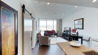 This contemporary single-floor residence showcases a thoughtfully designed open-concept layout that seamlessly connects the living, dining, and kitchen areas. A striking sliding barn door with a metal track serves as a stylish architectural accent, adding an industrial-modern touch to the interior. The living area is anchored by rich hardwood flooring and recessed lighting that illuminates the clean, white walls and high ceilings, creating a bright and airy atmosphere throughout. Floor-to-ceiling windows flood the space with abundant natural light while framing sweeping panoramic views of the surrounding cityscape and hills beyond — a truly standout feature of this residence. The living room comfortably accommodates a sectional sofa, accent chair, and a glass coffee table atop a light area rug, all oriented toward a media console and entertainment setup. The dining area, partially visible from the living space, connects directly to the kitchen, reinforcing the open and social nature of the floor plan. Bold red décor accents provide vibrant pops of color against the neutral palette of grays, taupes, and whites. The overall ambiance is polished, modern, and inviting — ideal for both relaxed everyday living and entertaining.