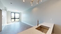 Located in Providence, Rhode Island, this contemporary single-floor unit showcases a thoughtfully designed open-concept layout with consistent herringbone-patterned flooring throughout, lending a refined and cohesive aesthetic to the entire space. The living and dining area is bathed in natural light from large, black-framed windows that frame outdoor views of the surrounding landscape, complemented by an elegant brass multi-arm pendant light fixture overhead.

The kitchen seamlessly integrates with the living area and is equipped with stainless steel appliances, including a full-size refrigerator, electric range, and built-in microwave. White upper cabinetry pairs with a bold navy blue kitchen island base, topped with a marble-veined quartz countertop and a brushed gold undermount sink with a pull-down faucet.

The bedroom offers a tranquil retreat with a large window providing outdoor views and access to a wall-mounted mini-split HVAC unit for individualized climate control. A sliding closet with built-in shelving and rod provides ample storage.

The bathroom is finished with modern sensibility, featuring a glass-enclosed walk-in shower with white tile surround, a wood-toned floating vanity with a white quartz countertop, an undermount sink, and a backlit mirror. Every detail throughout this space reflects clean, contemporary design.