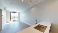 Located in Providence, Rhode Island, this contemporary single-floor unit showcases a thoughtfully designed open-concept layout with consistent herringbone-patterned flooring throughout, lending a refined and cohesive aesthetic to the entire space. The living and dining area is bathed in natural light from large, black-framed windows that frame outdoor views of the surrounding landscape, complemented by an elegant brass multi-arm pendant light fixture overhead.

The kitchen seamlessly integrates with the living area and is equipped with stainless steel appliances, including a full-size refrigerator, electric range, and built-in microwave. White upper cabinetry pairs with a bold navy blue kitchen island base, topped with a marble-veined quartz countertop and a brushed gold undermount sink with a pull-down faucet.

The bedroom offers a tranquil retreat with a large window providing outdoor views and access to a wall-mounted mini-split HVAC unit for individualized climate control. A sliding closet with built-in shelving and rod provides ample storage.

The bathroom is finished with modern sensibility, featuring a glass-enclosed walk-in shower with white tile surround, a wood-toned floating vanity with a white quartz countertop, an undermount sink, and a backlit mirror. Every detail throughout this space reflects clean, contemporary design.
