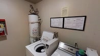 This dedicated laundry room is designed for practical, everyday use and contains all the essential utilities needed for on-site laundry. The room houses a top-loading washing machine and a matching dryer, both white and positioned side by side for easy access. A tall water heater is installed in the corner, supported on a raised platform with visible piping connections running along the wall above it. The walls feature posted instructional guides for the washer and dryer, displayed in a framed format and presented in multiple languages, ensuring clear and accessible guidance for all users. An additional notice is posted above the appliance guides, likely containing important usage or building-specific policies. The room's layout is compact and utilitarian, with neutral-toned walls and a straightforward design focused entirely on function. Electrical outlets are accessible along the wall near the appliances. Overall, this laundry room provides a self-contained, well-equipped space that meets standard residential laundry needs efficiently.