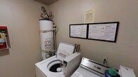 This dedicated laundry room is designed for practical, everyday use and contains all the essential utilities needed for on-site laundry. The room houses a top-loading washing machine and a matching dryer, both white and positioned side by side for easy access. A tall water heater is installed in the corner, supported on a raised platform with visible piping connections running along the wall above it. The walls feature posted instructional guides for the washer and dryer, displayed in a framed format and presented in multiple languages, ensuring clear and accessible guidance for all users. An additional notice is posted above the appliance guides, likely containing important usage or building-specific policies. The room's layout is compact and utilitarian, with neutral-toned walls and a straightforward design focused entirely on function. Electrical outlets are accessible along the wall near the appliances. Overall, this laundry room provides a self-contained, well-equipped space that meets standard residential laundry needs efficiently.