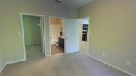 This single-floor space is designed with practical connectivity between its main living areas. The bedroom is finished with neutral-toned wall-to-wall carpeting that adds warmth underfoot, while soft sage-green walls create a calm, understated atmosphere throughout. The room benefits from notable ceiling height, lending a sense of openness to the space.

Two doorways lead directly from the bedroom — one opening into a private en-suite bathroom and the other into a walk-in closet fitted with shelving and hanging storage. The bathroom features wood-toned flooring, a dark vanity with sink, a toilet, and a bathtub with shower combination, all finished in clean, neutral tones with white fixtures and trim.

A second doorway from the bedroom provides a direct sightline into the kitchen, which is outfitted with black appliances, including a microwave and range, set against dark cabinetry. The layout promotes a seamless flow between the sleeping area and the kitchen. White door frames and baseboards throughout provide a crisp contrast to the muted wall tones, reinforcing a clean and cohesive interior aesthetic across the space.