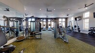 Located at 15155 Ollie Dr in Charlotte, NC, this fitness center is a thoughtfully designed, single-floor facility built to support a full range of workout routines. The space is divided into distinct functional zones, seamlessly blending strength training and cardiovascular areas within an open, well-lit environment.

The cardio section is positioned along a wall of large, floor-to-ceiling windows framed in dark trim, flooding the area with abundant natural light and offering views of the surrounding community. Multiple treadmills, elliptical machines, and stair climbers are arranged to take full advantage of this bright, airy setting.

The strength training area is equally well-appointed, featuring cable machines, a multi-station weight machine, a dedicated free weights section with a dumbbell rack, a power rack with barbecells, and various selectorized machines for targeted muscle groups. Wall-mounted mirrors expand the sense of space and allow users to monitor their form throughout their workouts.

The flooring transitions between patterned carpet tiles in the main zones and dark rubber flooring beneath the free weights, providing both comfort and safety. Overhead, ceiling fans and recessed lighting ensure consistent airflow and illumination throughout. Mounted flat-screen televisions and a clean, neutral color palette with contrasting dark accent walls contribute to a modern, motivating atmosphere.