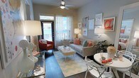 This single-floor space offers a thoughtfully designed interior that balances comfort and contemporary style. The living area serves as the centerpiece, featuring dark hardwood flooring that anchors the room with warmth and contrast. A ceiling fan overhead provides both function and a finishing architectural touch, while sheer curtain-draped windows allow abundant natural light to fill the space, creating an airy, inviting ambiance.

The living room is appointed with a neutral-toned sofa accented by coral and teal throw pillows, paired with a rust-colored accent chair that adds a bold pop of color. A distinctive faux-fur coffee table on acrylic legs serves as a stylish focal point atop a chevron-patterned area rug in soft blue and white tones. A carefully curated gallery wall featuring framed artwork in warm coral and neutral hues adds personality and depth to the space.

Adjacent to the living area, a compact dining nook features a round white table with sleek wire-frame chairs, perfect for casual dining. The overall color palette — soft grays, whites, and warm coral accents — creates a cohesive and vibrant aesthetic throughout. The space makes excellent use of its layout, maximizing functionality while maintaining an open, welcoming feel suited for everyday living and entertaining alike.