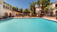 Located at 1001 Starbuck St in Fullerton, California, this outdoor amenity space centers around a generously sized, crystal-clear swimming pool that serves as the focal point of the community. The pool is framed by a wide, tile-edged deck outfitted with rows of lounge chairs and shade umbrellas, providing ample space for sunbathing and relaxation under the Southern California sun.

The surrounding architecture features a Mediterranean-inspired clubhouse building with tall arched windows, smooth stucco facades, and elegant proportions that lend the area a refined, resort-like atmosphere. A separate lounge patio area offers wicker seating with blue cushions and a patio umbrella, nestled alongside a stone-accented retaining wall — ideal for casual outdoor gatherings.

Mature palm trees, tropical plantings, and lush greenery border the pool deck on multiple sides, creating a sense of privacy and a vibrant, verdant backdrop. The overall layout, as visible in the aerial scan, reveals a well-organized outdoor environment with distinct zones for swimming, lounging, and socializing. The combination of thoughtful landscaping, quality furnishings, and architectural detail makes this outdoor space a standout community amenity within the Fullerton area.