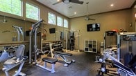 This single-floor fitness center is thoughtfully designed to support a full range of workout routines. The room features elevated ceilings with clerestory windows along the upper walls, allowing ample natural light to filter in while maintaining privacy. Two ceiling fans are mounted overhead, ensuring air circulation throughout workouts.

The space is outfitted with a robust selection of strength training equipment, including a multi-function cable machine, an adjustable weight bench, and a well-stocked dumbbell rack spanning a wide range of weights. A cable crossover tower anchors the center of the room, while additional free weights and specialty equipment are positioned along the perimeter.

Large wall mirrors span one side of the room, visually expanding the space and allowing users to monitor their form during exercise. Two wall-mounted flat-screen televisions provide entertainment options. The flooring consists of durable rubber gym matting, designed to absorb impact and protect equipment. An open-cube storage unit offers space for personal items or accessories. The overall ambiance is practical and functional, with neutral wall tones and recessed lighting complementing the natural light from the windows.