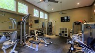 This single-floor fitness center is thoughtfully designed to support a full range of workout routines. The room features elevated ceilings with clerestory windows along the upper walls, allowing ample natural light to filter in while maintaining privacy. Two ceiling fans are mounted overhead, ensuring air circulation throughout workouts.

The space is outfitted with a robust selection of strength training equipment, including a multi-function cable machine, an adjustable weight bench, and a well-stocked dumbbell rack spanning a wide range of weights. A cable crossover tower anchors the center of the room, while additional free weights and specialty equipment are positioned along the perimeter.

Large wall mirrors span one side of the room, visually expanding the space and allowing users to monitor their form during exercise. Two wall-mounted flat-screen televisions provide entertainment options. The flooring consists of durable rubber gym matting, designed to absorb impact and protect equipment. An open-cube storage unit offers space for personal items or accessories. The overall ambiance is practical and functional, with neutral wall tones and recessed lighting complementing the natural light from the windows.