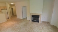 This single-floor space offers a straightforward and functional layout designed for comfortable everyday living. The main living area is a standout feature, highlighted by a tile-surround fireplace with a black metal insert that serves as a natural focal point of the room. The raised tile hearth and clean surround give the fireplace a classic, understated look that complements the neutral interior palette. Wall-to-wall carpeting in a warm beige tone extends throughout the living space, adding softness underfoot and contributing to the overall cozy ambiance. The walls are finished in a light, muted green-white tone that keeps the space feeling bright and airy. A doorway visible from the living area leads to an adjacent room, suggesting a connected and accessible floor plan. The space benefits from an open feel with generous room dimensions that allow for flexible furniture arrangements. The overall aesthetic is clean and neutral, providing an excellent canvas for personalization. With its combination of practical finishes, a welcoming fireplace, and a cohesive color scheme, this space offers a comfortable and inviting environment suited to a variety of lifestyles.