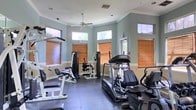 This fitness center is thoughtfully designed to support a full range of workout routines within a clean, comfortable single-floor space. The room features elegant crown molding and a neutral sage-green color palette along the upper walls, complemented by crisp white wainscoting below, giving the space a polished, well-maintained appearance. A ceiling fan mounted at the center of the room aids in air circulation during workouts, contributing to a pleasant exercise environment. Natural light filters in through multiple transom-style windows fitted with warm wood-toned blinds, offering a bright yet controllable atmosphere. The flooring consists of durable dark rubber matting, practical for heavy equipment use and easy maintenance. The equipment selection is diverse, including a Landice treadmill, a multi-station cable and weight machine, a recumbent-style elliptical or bike, and various strength-training benches and attachments. The layout is organized to allow users to navigate between stations with ease, making the space functional for both cardio and resistance training. Overall, this fitness center combines practical amenities with tasteful architectural details to deliver an inviting and fully operational workout facility.