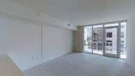 This single-floor space offers a clean, contemporary aesthetic defined by minimalist design and an abundance of natural light. The main living area is characterized by crisp white walls and smooth ceilings with recessed ventilation, creating an open and airy atmosphere that feels expansive and uncluttered. Light-toned hardwood flooring runs throughout, lending warmth and continuity to the space while complementing the bright, neutral palette.

A striking floor-to-ceiling glass sliding door serves as a focal point, flooding the interior with natural daylight and providing direct access to an outdoor balcony. The balcony overlooks an urban cityscape, offering views of the surrounding buildings and skyline. This seamless indoor-outdoor connection enhances the sense of openness and makes the space feel larger than its footprint suggests.

A paneled interior door provides access to an adjacent room, suggesting a thoughtful and functional layout. The overall design sensibility is modern and understated, with clean lines and a neutral color scheme that offers a versatile canvas for a variety of uses. The combination of ample natural light, stylish flooring, and outdoor access makes this a well-appointed and inviting space suited to contemporary living.