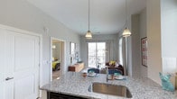 This 1-bedroom, 1-bathroom residence offers a thoughtfully designed open-concept layout that seamlessly connects the kitchen, dining, and living areas. The kitchen is a standout feature, boasting granite countertops with a speckled finish, a stainless steel undermount sink, and stylish pendant lighting that adds a modern touch overhead. The kitchen island serves as a natural gathering point, providing additional prep space and casual seating. The living area is bright and welcoming, benefiting from a large sliding glass door that opens to a private balcony, allowing ample natural light to fill the space and offering views of the surrounding community. Neutral gray walls and light flooring throughout create a clean, contemporary aesthetic. The bedroom, visible through an open doorway, features carpeted flooring and continues the light, neutral color palette. With its modern finishes, functional open floor plan, and access to outdoor space, this residence provides a comfortable and stylish living environment suited for everyday life.