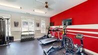 Located at 5003 Winster Dr in Winston-Salem, NC, this fitness center offers a compact yet thoroughly equipped workout space spread across a single floor. The room makes a strong visual impression with a bold red accent wall accented by white horizontal stripes, providing an energetic atmosphere conducive to exercise. Two flat-screen TVs are mounted on the accent wall, allowing members to stay entertained during cardio sessions.

The space is outfitted with multiple treadmills and elliptical trainers positioned along the feature wall, while a multi-station cable machine and a dedicated free weight rack with a full dumbbell set line the opposing side. A weight bench with a barbell rack adds further strength-training capability. Full-length mirrors along one wall amplify the sense of space and allow users to monitor their form during workouts.

Natural light streams in through several large windows fitted with elegant valances, offering views of lush greenery outside. Durable rubber flooring covers the entire area, providing both safety and comfort underfoot. Ceiling fans and track lighting ensure the space remains comfortable and well-lit at all times. A glass-panel entry door connects the fitness center to the broader property, reinforcing its accessibility and community-oriented design.