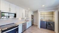 Located at 2 Victorian Court in Holtsville, NY, this two-floor townhouse at Fairfield Townhouses offers a thoughtfully updated interior with a clean, modern aesthetic throughout. Light-toned hardwood-style flooring flows consistently across both levels, complemented by neutral gray walls and crisp white trim that create a bright, cohesive atmosphere.

The main living area is generously proportioned, featuring crown molding, recessed lighting, and a ceiling fan. A sliding glass door opens directly onto an outdoor deck, inviting natural light and providing convenient access to the exterior. Baseboard heating runs along the perimeter, and a wall-mounted air conditioning unit ensures year-round comfort.

The kitchen is a standout feature, outfitted with white shaker-style cabinetry—including glass-front upper cabinets—and a marble-look tile backsplash. Light quartz countertops and a stainless steel undermount sink are paired with a full suite of stainless steel appliances, including a slide-in range, over-the-range microwave, dishwasher, and French door refrigerator. Adjacent to the kitchen is a dedicated laundry area with a full-size washer and dryer and built-in shelving for storage.

The bathroom features a tub-shower combination enclosed by a sleek glass panel, with white subway tile surround and built-in corner shelving. A staircase connects both floors, completing this well-appointed townhouse.