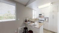 This single-story residence offers a thoughtfully arranged layout with distinct living, sleeping, kitchen, and bathroom areas. The open living room is finished with light gray wood-look flooring and features a large sliding glass door fitted with vertical blinds, allowing generous natural light to fill the space and providing access to an outdoor area. A ceiling fan adds a functional touch overhead, while a textured ceiling adds subtle architectural character.

The kitchen is efficiently designed around a peninsula that doubles as a casual dining bar, with seating provided by dark bar stools. Granite-look countertops, white cabinetry, a tile backsplash, and wood-look flooring create a clean, cohesive aesthetic. The kitchen is equipped with a gas range, over-the-range microwave, and ample cabinet storage. Multiple windows supply natural light throughout the cooking area.

The bedroom is carpeted and spacious, featuring neutral taupe walls, a textured ceiling, and a window with horizontal blinds. A notable convenience is the in-unit stacked washer and dryer, neatly housed in a dedicated closet within the room.

The bathroom includes a white soaking tub with shower, a granite-look vanity countertop with white cabinetry, a large mirror, and wood-look flooring, completing the unit's consistent and practical design.
