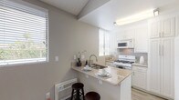 This single-story residence offers a thoughtfully arranged layout with distinct living, sleeping, kitchen, and bathroom areas. The open living room is finished with light gray wood-look flooring and features a large sliding glass door fitted with vertical blinds, allowing generous natural light to fill the space and providing access to an outdoor area. A ceiling fan adds a functional touch overhead, while a textured ceiling adds subtle architectural character.

The kitchen is efficiently designed around a peninsula that doubles as a casual dining bar, with seating provided by dark bar stools. Granite-look countertops, white cabinetry, a tile backsplash, and wood-look flooring create a clean, cohesive aesthetic. The kitchen is equipped with a gas range, over-the-range microwave, and ample cabinet storage. Multiple windows supply natural light throughout the cooking area.

The bedroom is carpeted and spacious, featuring neutral taupe walls, a textured ceiling, and a window with horizontal blinds. A notable convenience is the in-unit stacked washer and dryer, neatly housed in a dedicated closet within the room.

The bathroom includes a white soaking tub with shower, a granite-look vanity countertop with white cabinetry, a large mirror, and wood-look flooring, completing the unit's consistent and practical design.