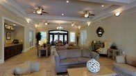 This inviting single-floor space exudes a refined yet comfortable ambiance, thoughtfully designed to welcome guests and encourage relaxation. The interior is anchored by an impressive coffered ceiling adorned with multiple ceiling fans and recessed lighting, lending both architectural character and practical comfort to the room. A central chandelier adds a touch of elegance, complementing the warm, neutral color palette that flows throughout the space. Large French doors and oversized windows line the walls, flooding the interior with natural light while offering views of the lush greenery outside. The open layout is furnished with plush seating arrangements, including a generous sofa and coordinating armchairs grouped on area rugs, creating distinct yet connected conversation areas. Decorative accents such as sculptural vases, potted plants, and a distinctive round mirror contribute to the space's polished aesthetic. Stone tile flooring ties the room together with a sense of understated sophistication. Whether used for socializing or quiet relaxation, this space masterfully balances style and function, offering an atmosphere that is both upscale and approachable.