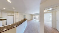 This single-floor residence is designed with an open, functional layout that connects the kitchen and living areas seamlessly. The kitchen is a standout feature, equipped with white cabinetry extending to the ceiling, dark granite countertops, wood-look flooring, and white appliances including a range, microwave, and refrigerator. Recessed lighting illuminates the space effectively. A peninsula with a matching granite surface and an integrated sink separates the kitchen from the adjacent living area, providing both additional workspace and a casual dining option.

The living area is generously sized, finished with plush neutral carpet, and anchored by a sliding glass door that opens to a private balcony. The balcony offers views of the surrounding community and allows natural light to flood the interior. Additional bedrooms are carpeted and well-proportioned, each featuring windows with blinds that bring in natural light. One bedroom also provides direct balcony access via its own sliding glass door.

The bathroom features wood-look flooring and recessed lighting in a clean, neutral palette. Throughout the residence, the consistent use of soft white walls, neutral carpeting, and warm wood-look flooring creates a cohesive and bright ambiance.